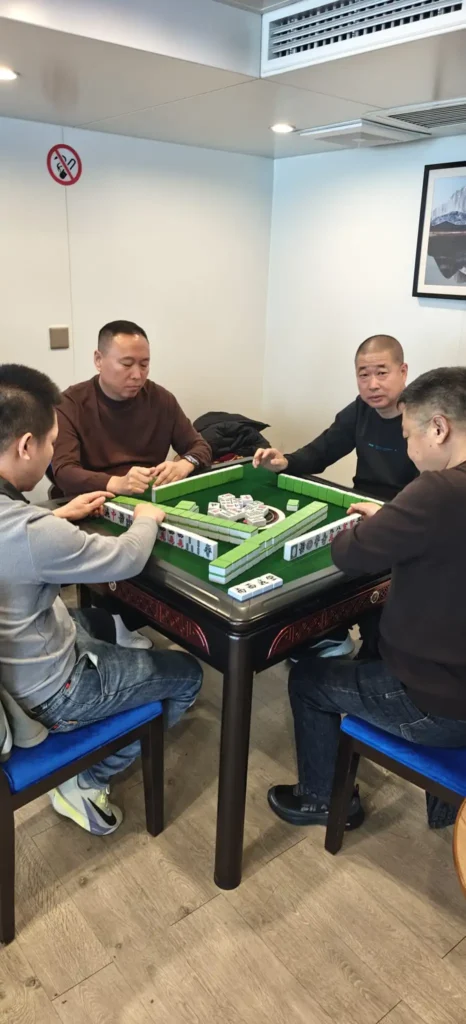 Cuatro hombres están sentados alrededor de una mesa de mahjong, jugando una partida. La mesa está cubierta de fichas de mahjong. La habitación, que evoca el ambiente de Chongqing, tiene las paredes blancas, un cuadro enmarcado y un cartel de prohibido fumar.