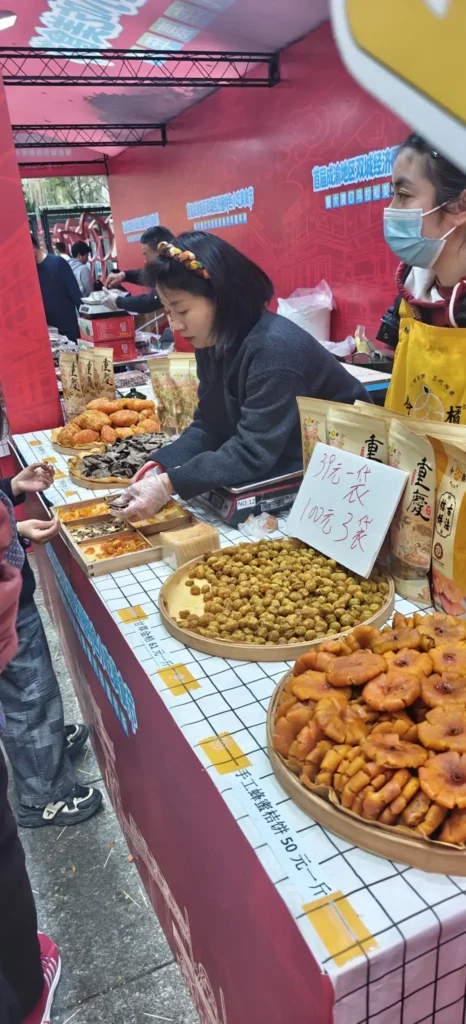 Una mujer detrás de un puesto de un mercado de Shanghái en el que se exponen diversos aperitivos fritos y productos envasados. Sirve comida a los clientes mientras otra vendedora con delantal amarillo observa. Un cartel escrito a mano indica los precios en chino.
