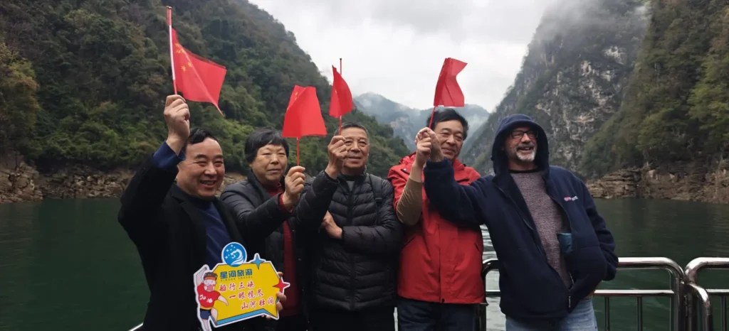 Cinco adultos en una barca por el Río Azul, cerca de Chongqing, con banderitas chinas en la mano y sonriendo, con el agua verde y las montañas rocosas y boscosas de fondo. Una persona sostiene un cartel de colores con texto en chino.