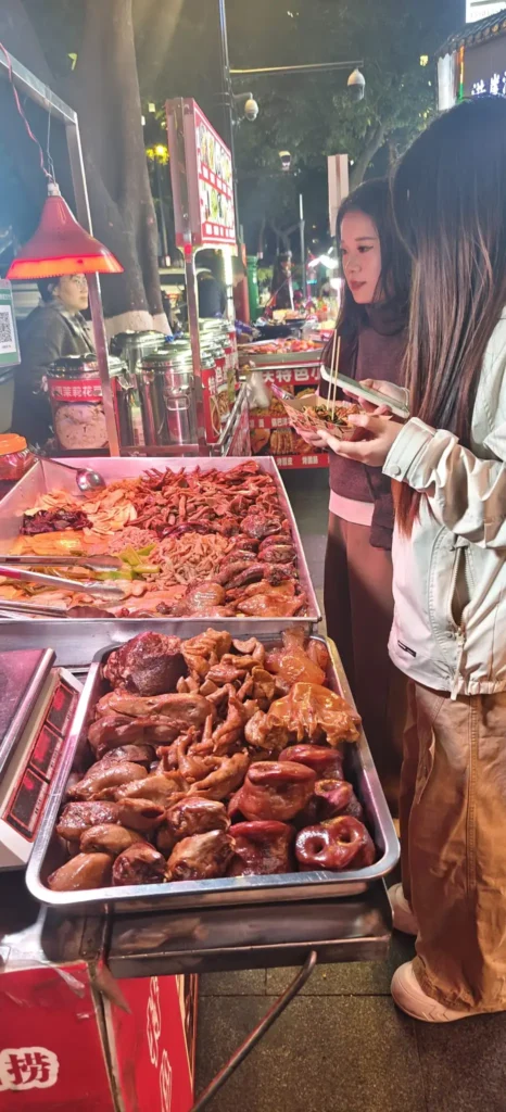 Dos mujeres en un puesto de comida callejera de Chongqing, iluminado por la noche, seleccionan con palillos varias carnes y despojos de bandejas metálicas. El bullicioso puesto exhibe una variedad de platos preparados típicos de la vibrante escena gastronómica de la ciudad.