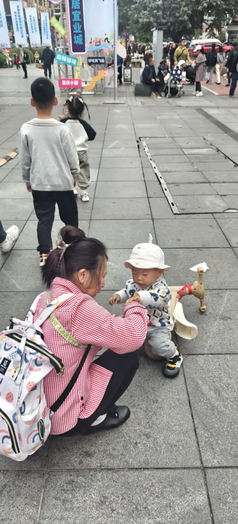 Una mujer se agacha junto a un niño pequeño con gorro blanco y chaqueta estampada en una zona exterior pavimentada de Chongqing. El niño está cerca de un caballito de madera, y al fondo se ve a otros niños y adultos.