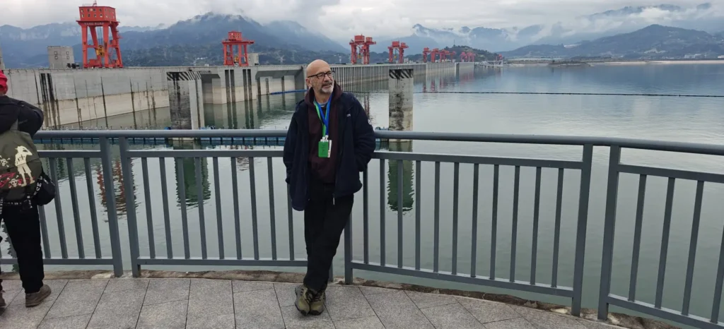 Una persona frente a una barandilla metálica con vistas a una gran presa en el Río Azul, con estructuras rojas y aguas tranquilas; montañas y nubes se alzan al fondo, cerca de Chongqing.