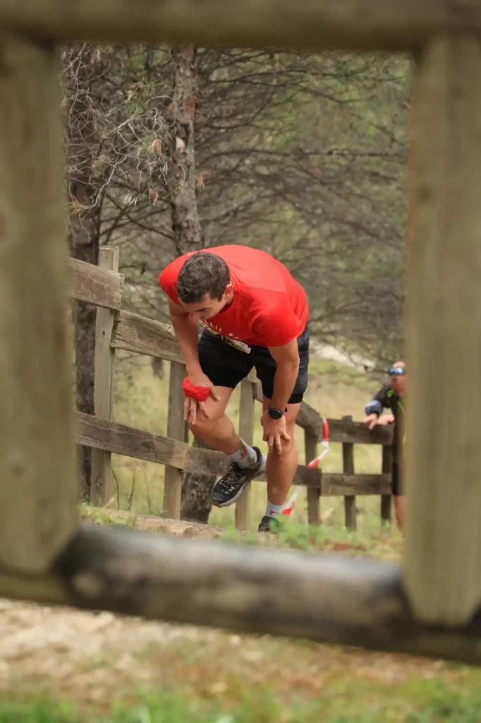 Un hombre con camiseta roja y pantalones cortos negros se inclina hacia delante, agarrándose la rodilla en Rivas Trail Natura, cerca de una valla de madera. A pesar de la ligera lluvia, al fondo se ven otros corredores y árboles.