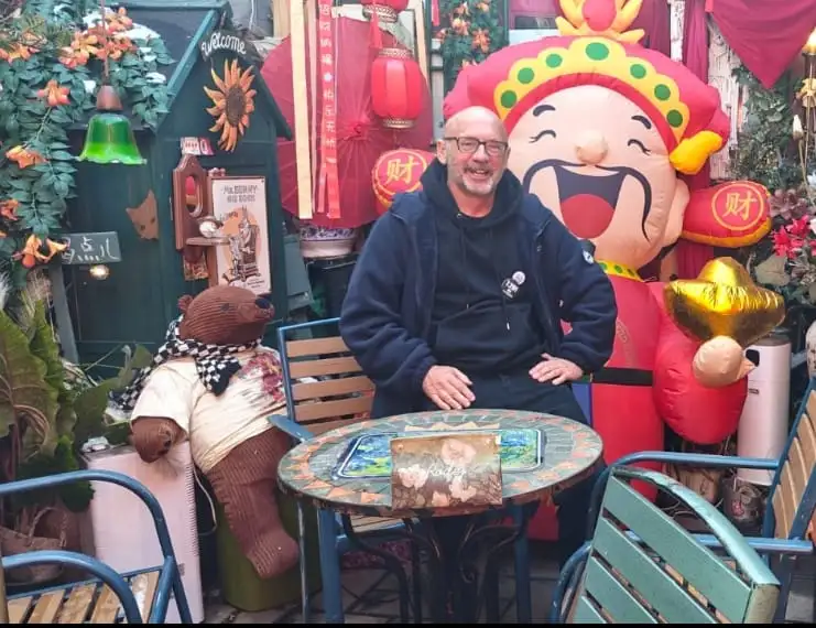 Un hombre con gafas y sudadera oscura se sienta en una colorida mesa de mosaico al aire libre durante la Semana 1, rodeado de decoraciones festivas, un gran personaje hinchable, un osito de peluche, plantas y farolillos rojos-memorias por China como bajista afortunado.