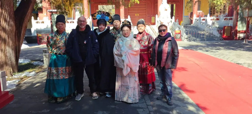Un grupo de ocho personas posa al aire libre; algunas llevan trajes tradicionales chinos y otras ropa moderna de invierno. Están de pie en un camino de piedra junto a una alfombra roja, captando Memorias por China junto a un edificio histórico y árboles al fondo.
