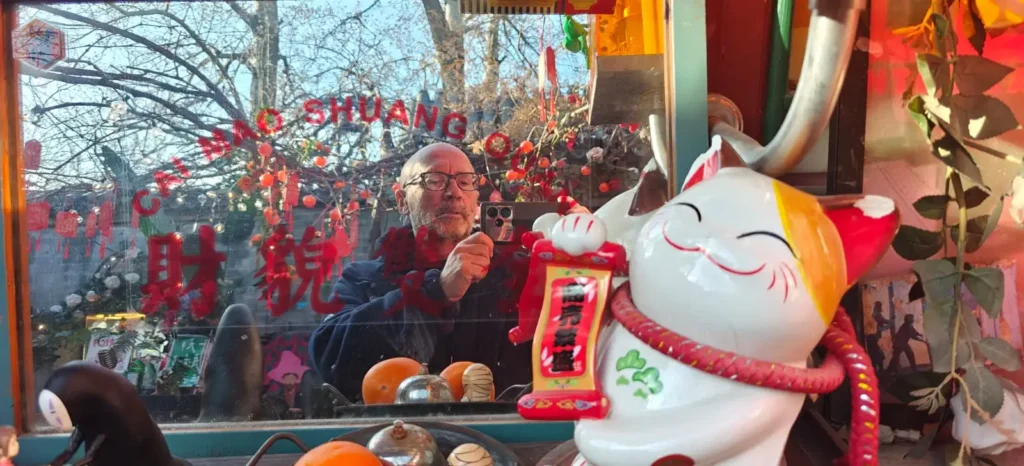 Una persona hace una foto a través de una ventana decorada con caracteres y adornos chinos rojos, que evocan recuerdos de China. Una gran figura de Maneki-neko se sitúa en primer plano, con reflejos y diversos objetos dentro y fuera.