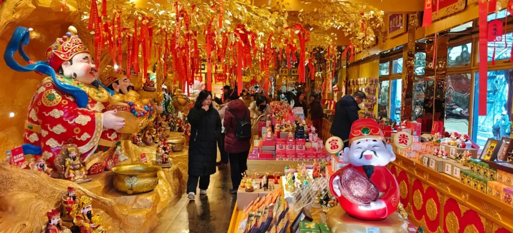 Una tienda china decorada con colores vivos está repleta de estatuas doradas, farolillos rojos y adornos festivos. Varias personas hojean estanterías repletas de diversos artículos, mientras que grandes estatuillas y adornos colgantes ocupan un lugar destacado.