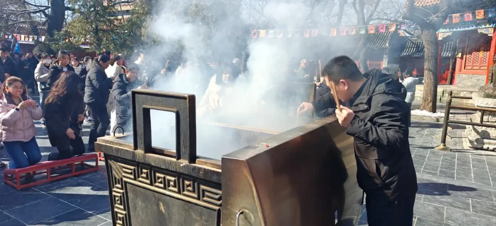 Un hombre de pie frente a un gran incensario con humo ascendente, sosteniendo varitas de incienso, mientras varias personas se arrodillan y rezan en un concurrido templo al aire libre en China.