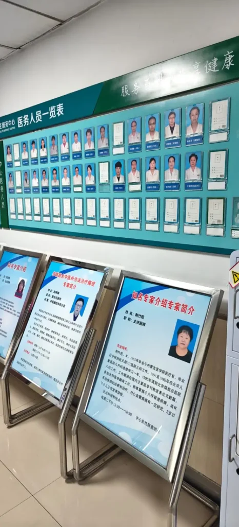 Un expositor mural en China muestra fotos y nombres de personal médico en uniforme, dispuestos en filas bajo un encabezamiento verde. Delante, tres tableros sobre atriles presentan hojas informativas más grandes con fotos y textos que recuerdan su servicio.