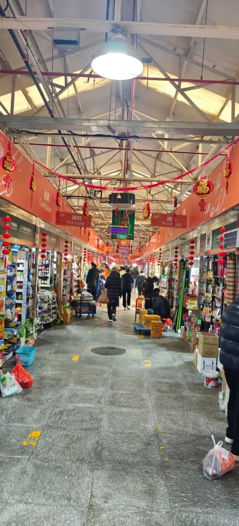 Mercado cubierto en China con decoraciones rojas y farolillos colgantes, gente comprando en puestos llenos de mercancías y comestibles, mientras un bajista toca cerca, creando nuevos recuerdos bajo el techo de claraboya.