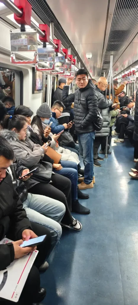 Pasajeros con chaquetas de invierno, algunos sumidos en sus memorias, se sientan y permanecen de pie en el interior de un tren de metro en China. La mayoría mira sus teléfonos. El tren tiene el suelo azul, las paredes plateadas y asideros rojos colgando del techo.
