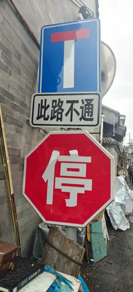 Una señal de calle indica un callejón sin salida encima de una señal de stop octogonal roja con caracteres chinos, montada en un poste junto a una pared de ladrillo gris en un callejón al aire libre en China, con varios objetos apilados cerca.