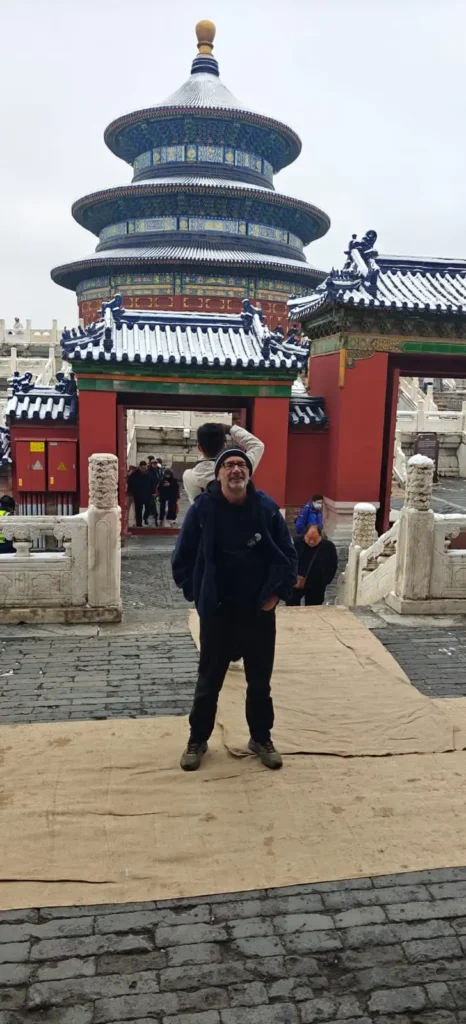 Una persona sonríe delante de la Sala de Oración por las Buenas Cosechas del Templo del Cielo de Pekín, China, en un día nublado. La nieve espolvorea el techo y el suelo mientras crean recuerdos, mientras otros visitantes exploran el fondo.