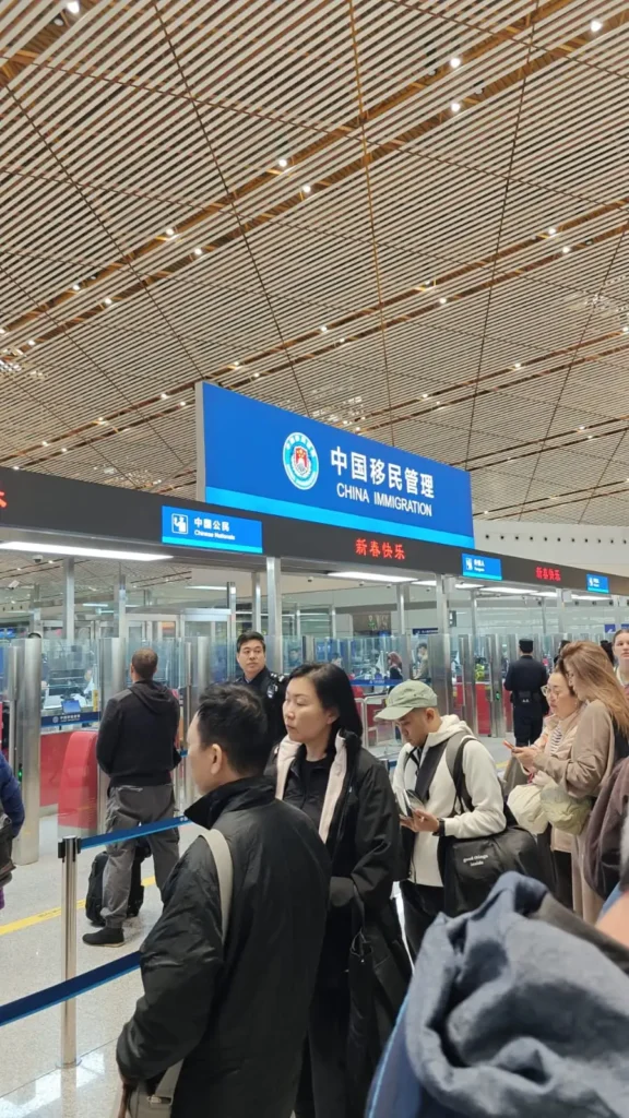 Un grupo de personas hace cola en un puesto de control de inmigración de China, quizás creando nuevos recuerdos mientras esperan bajo el cartel azul. La zona, bien iluminada, tiene barreras de cristal y mostradores a la vista, lo que contribuye al ambiente ordenado.