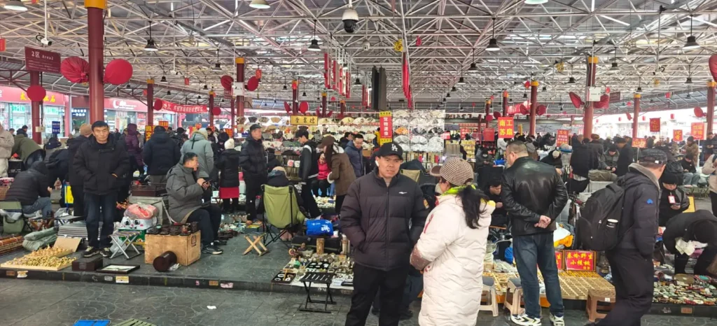 Un ajetreado mercado cubierto en China, con mucha gente curioseando y vendiendo diversos artículos expuestos en el suelo y en mesas. El mercado está bien iluminado, con techo metálico, banderolas rojas colgadas por todas partes, y alberga innumerables recuerdos para los visitantes.