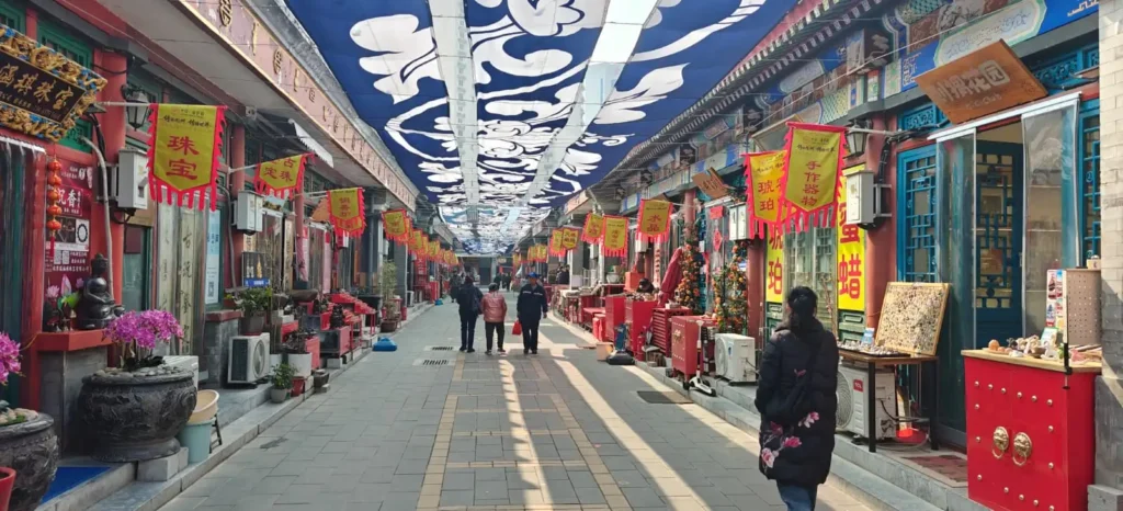 Una calle peatonal de China repleta de tiendas exhibe pancartas rojas y amarillas. Varias personas caminan por la acera, recogiendo recuerdos bajo telas estampadas en azul y blanco, mientras la luz del sol proyecta sombras sobre la escena.