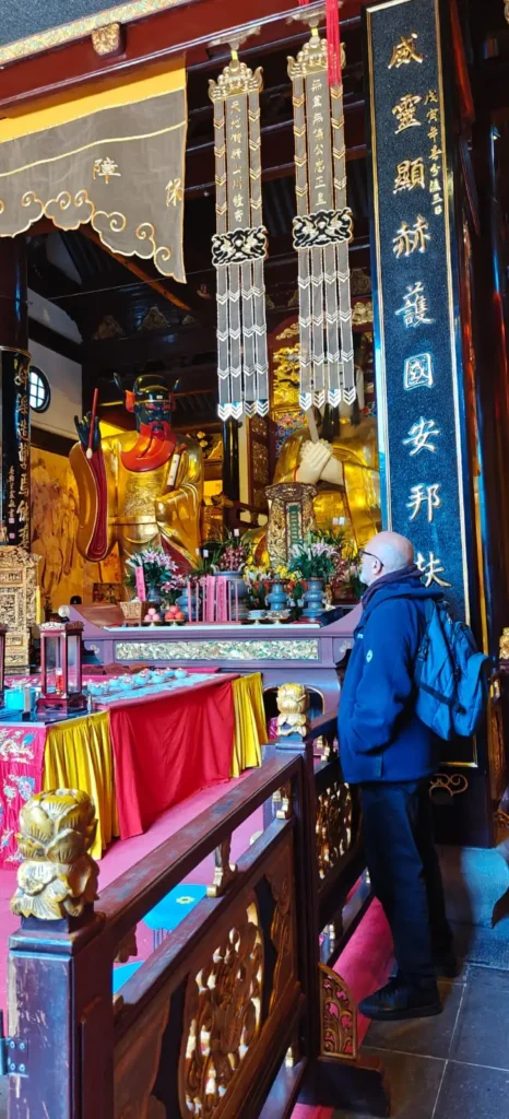 Una persona con chaqueta azul y mochila se encuentra en el interior de un templo, contemplando un altar ornamentado con adornos dorados, estatuas, flores, velas rojas y caligrafía china: recuerdos por China durante la Semana 1.