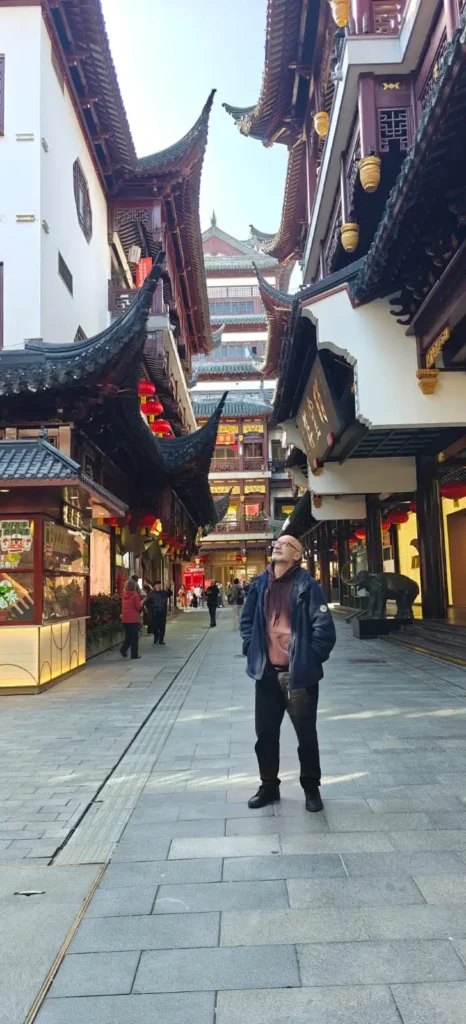 Un hombre se encuentra en medio de una calle tradicional china con tejados ornamentados, rodeado de tiendas y farolillos rojos. La brillante luz del día resalta la detallada arquitectura: comienza su viaje: Memorias por China, Semana 1.