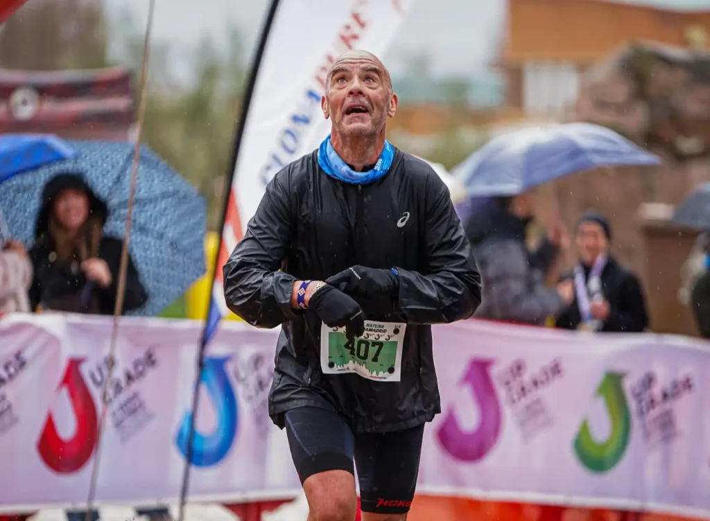 Un hombre con chaqueta negra y pantalón corto cruza la línea de meta de la carrera Rivas Trail Natura mirando hacia arriba. Espectadores con paraguas se sitúan al fondo, cerca de pancartas, mientras cae la lluvia, destacando a los corredores entregados a pesar del tiempo lluvioso.
