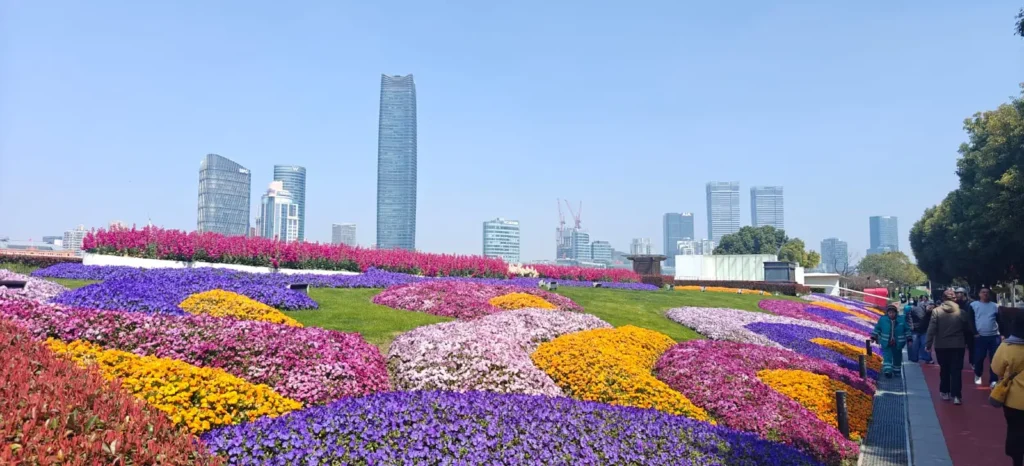 Paisaje urbano en Pekín con modernos rascacielos al fondo y un gran parterre en suave pendiente en primer plano, con coloridas manchas de flores moradas, amarillas, rojas y rosas. La gente pasea por el camino de la derecha.