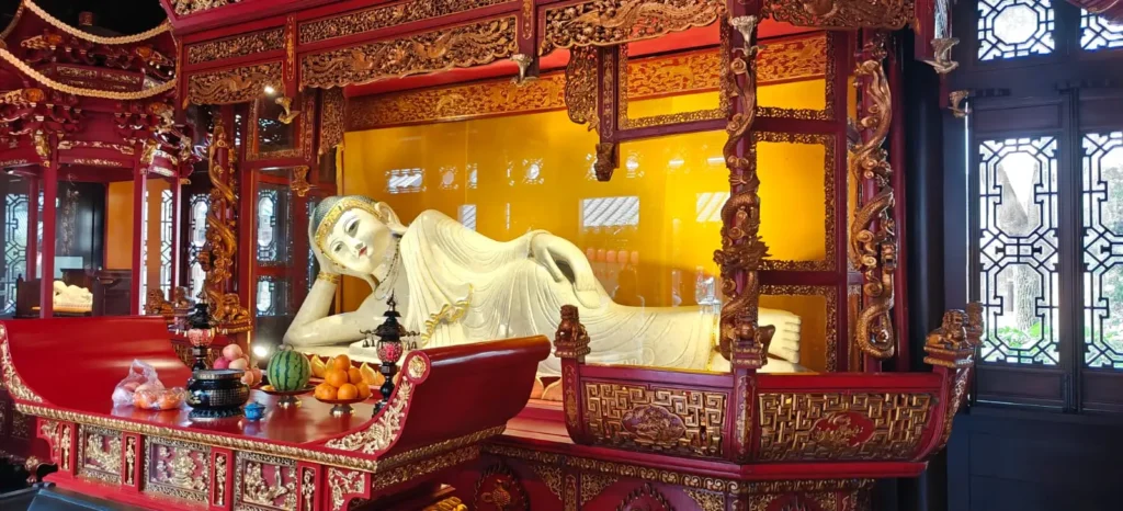 Una estatua blanca de Buda reclinado se exhibe en un ornamentado santuario rojo y dorado, que recuerda la grandiosidad de Pekín (China). Ofrendas de fruta y velas descansan sobre una mesa ante intrincadas tallas y decoraciones que rodean la escena.