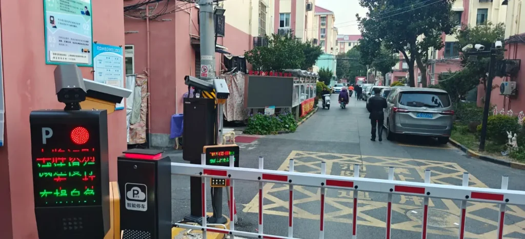 Entrada de una calle residencial en Pekín (China), con una barrera, máquinas de pago por aparcamiento y una señal de semáforo en rojo. Varios coches aparcan cerca mientras la gente entra en la zona residencial, lo que refleja la influencia de la seguridad occidental en la planificación urbana.