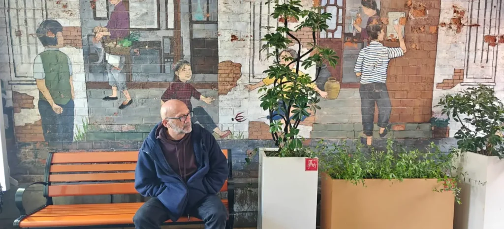 Un hombre con barba está sentado en un banco naranja en el interior de Pekín, mirando a su derecha. Detrás de él hay un mural que representa a personas realizando actividades cotidianas, con dos plantas en macetas delante del mural.