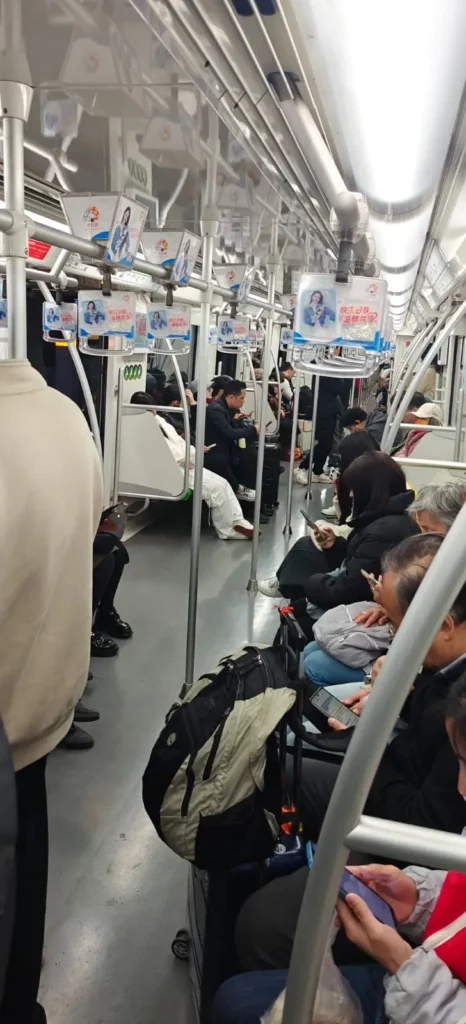 Pasajeros sentados y de pie en el interior de un tren subterráneo en Pekín. La mayoría mira el móvil o descansa, con chaquetas y bolsos cerca. Encima hay anuncios y pasamanos. El ambiente tranquilo y silencioso refleja la vida cotidiana en China.