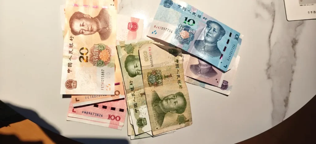 Sobre una superficie blanca y gris se extienden billetes chinos de diversas denominaciones, como 100, 20, 10 y 5 yuanes. En la mayoría de los billetes aparece un retrato de Mao Zedong, símbolo frecuente en toda China y en su capital, Pekín.