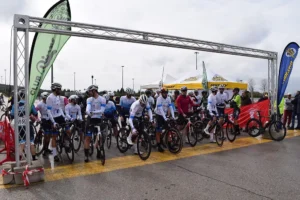 Un grupo de ciclistas con maillots a juego espera en la línea de salida de una carrera de carretera, listos para el homenaje a Rivas y Carlos Cubillo. Bicicletas alineadas, pancartas, carpas y autoridades visibles al fondo bajo un cielo nublado.