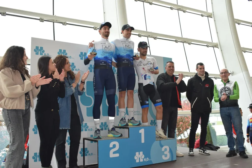 Tres ciclistas, entre ellos Carlos Cubillo, suben al podio de los vencedores, dos de los cuales comparten el escalón más alto con sus equipaciones de competición. En las inmediaciones, la gente aplaude y sonríe durante este acto en Rivas, celebrado como homenaje con grandes ventanales al fondo.