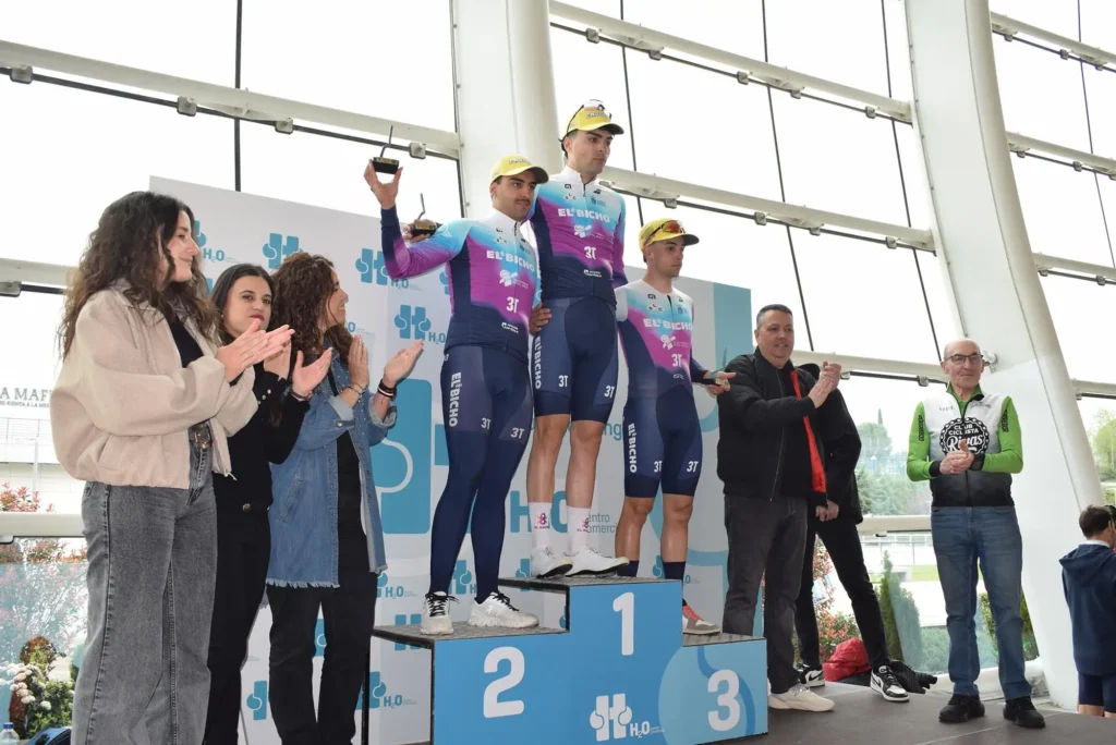 Tres ciclistas con uniformes a juego y gorras amarillas suben a un podio de ganadores, sosteniendo trofeos durante un homenaje en Rivas. Varias personas junto al podio aplauden y sonríen. El acto parece celebrarse en un interior con grandes ventanales al fondo.