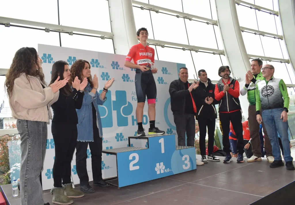 Un ciclista con maillot rojo sube a lo más alto del podio sosteniendo un trofeo, mientras la gente en el escenario a su alrededor aplaude durante un homenaje a Carlos Cubillo en el interior de un edificio con grandes ventanales en Rivas.