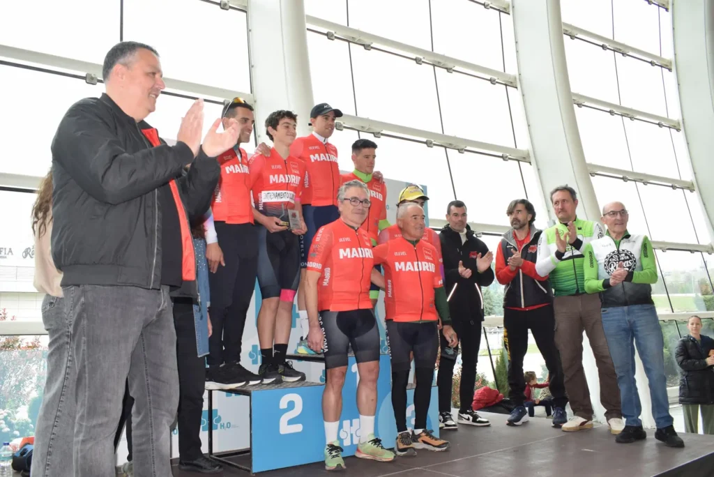 Un grupo de ciclistas con maillots rojos a juego suben a un podio en el interior de un edificio para celebrar el homenaje a Carlos Cubillo en Rivas. Varias personas aplauden y posan para una foto mientras un ciclista sostiene un trofeo.