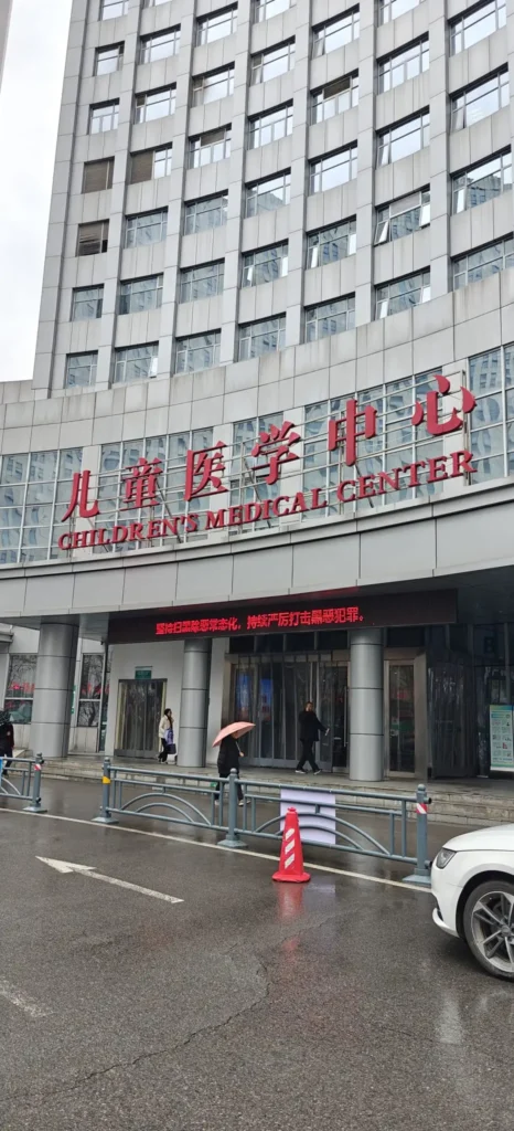 La entrada de un edificio de varios pisos en Chongqing con grandes letras rojas que dicen Childrens Medical Center sobre las puertas; la gente está de pie fuera bajo paraguas en un día lluvioso.