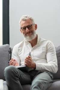 Un hombre de pelo y barba canosos, con gafas y camisa blanca de botones, sentado en un sofá gris con un cuaderno y un bolígrafo en la mano, reflexiona sobre la jubilación anticipada para los trabajadores con más de 40 años cotizados y mira a la cámara con una leve sonrisa.