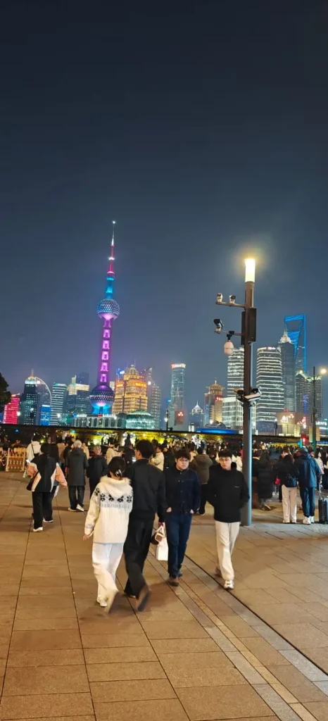 La gente camina por un paseo marítimo de noche en China, con el horizonte de Shanghái -incluida la Torre de la Perla Oriental iluminada- brillando a sus espaldas. Las farolas y las luces de la ciudad resaltan el vibrante paisaje urbano, reflejando una China moderna más allá de Pekín.