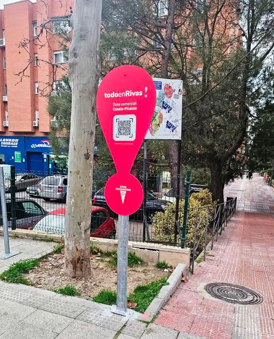 Un cartel rojo en forma de signo de exclamación con un código QR y el texto todoenRivas se encuentra en una acera cerca de coches aparcados, un árbol y un edificio de ladrillo en una zona urbana.