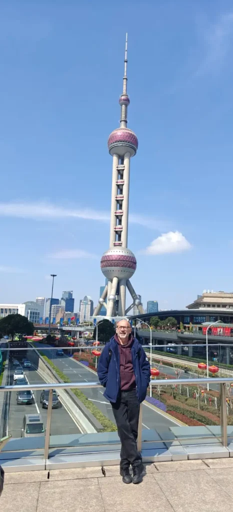 Una persona sonríe delante de la Torre de la Perla Oriental en Shanghai (China), con un cielo azul despejado, edificios urbanos y una calle muy transitada con vehículos, que retrata la China moderna más allá de Pekín o los temas de seguridad occidental.