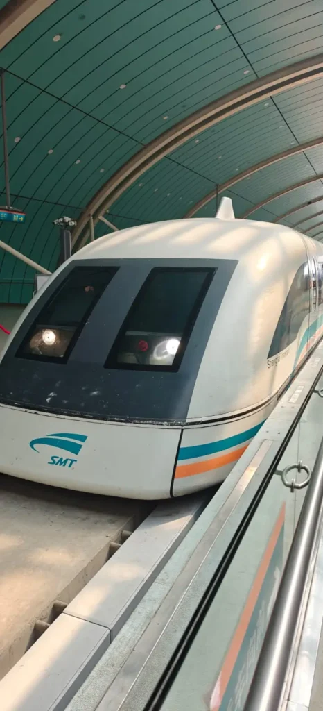 Un tren de levitación magnética SMT blanco con franjas verde azulado y naranja se detiene en un andén dentro de una moderna estación de techo curvo con paneles verdes en Pekín. Los faros del tren están encendidos y las barreras de cristal garantizan la seguridad occidental a lo largo del borde del andén.