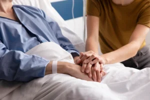 Una persona en una cama de hospital con una bata azul y una muñequera se da la mano con otra persona de pie junto a la cama, ofreciéndole apoyo. Solo se ven la parte superior del cuerpo y las manos.