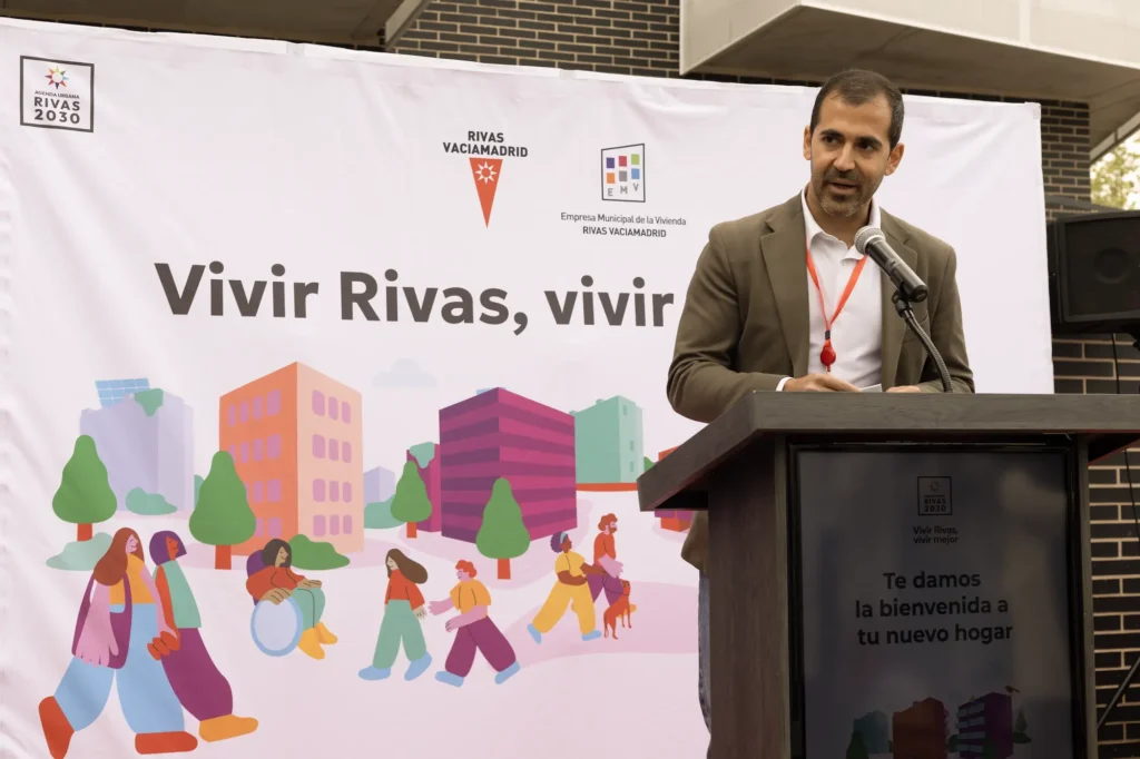 Un hombre trajeado habla por un micrófono. Detrás de él, una pancarta del Ayuntamiento de Rivas con coloridas ilustraciones de edificios y personas destaca iniciativas como las viviendas públicas y el alquiler para jóvenes en este acto al aire libre.