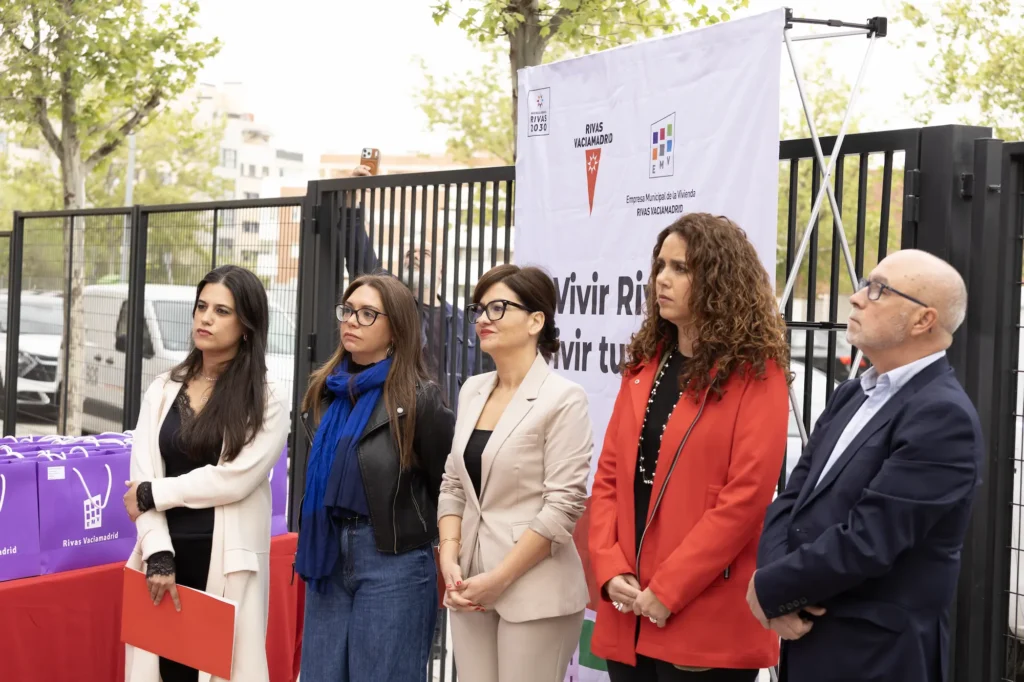 Cinco adultos en fila al aire libre, cerca de una valla metálica, mientras tres mujeres y un hombre escuchan atentamente un acto del Ayuntamiento de Rivas sobre viviendas públicas. Cerca hay una pancarta y una mesa con bolsas moradas, con árboles y edificios al fondo.