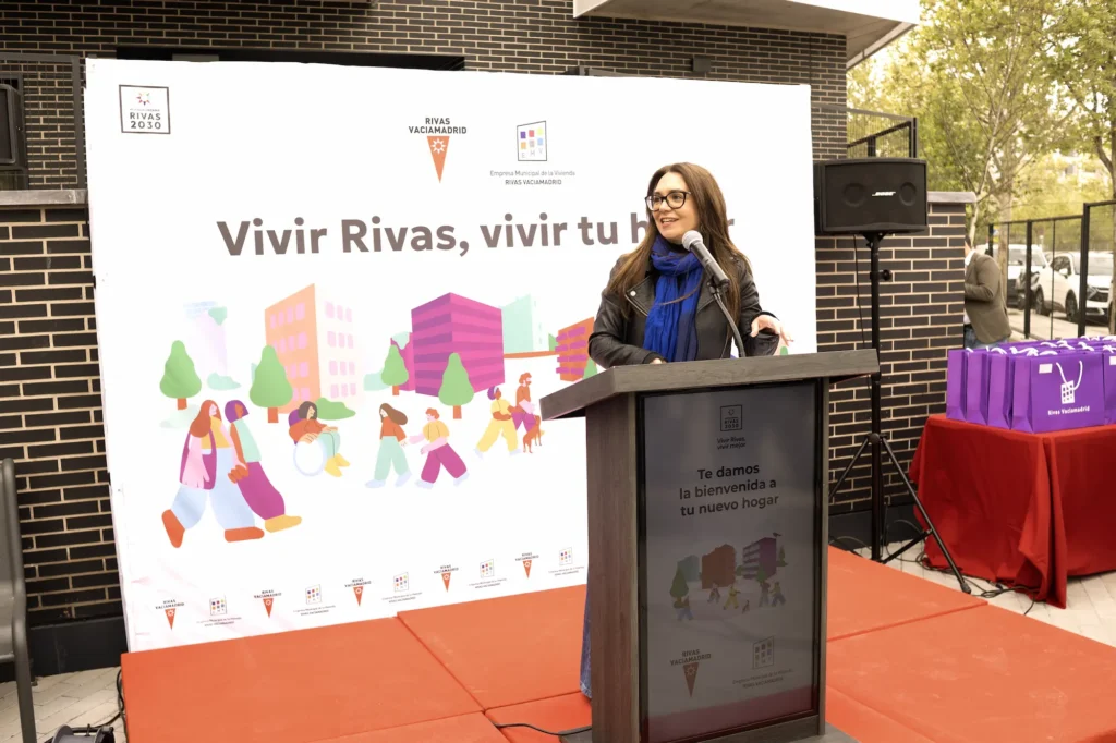 Una mujer habla ante un micrófono en un acto al aire libre. Detrás de ella, una pancarta con coloridas ilustraciones que reza Vivir Rivas, vivir tu hogar destaca el compromiso del Ayuntamiento de Rivas con las viviendas públicas y el alquiler para jóvenes.