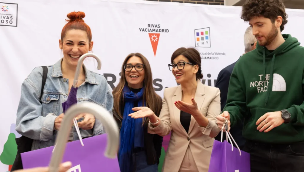 Cuatro personas se sit&uacute;an frente a una pancarta de Rivas, sonrientes y con bolsas moradas en las manos. Dos mujeres en el centro gesticulan con las manos, mientras un hombre con sudadera verde mira un objeto relacionado con alquiler para j&oacute;venes. En primer plano se ve el mango de un paraguas.