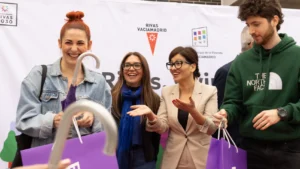 Cuatro personas se sitúan frente a una pancarta de Rivas, sonrientes y con bolsas moradas en las manos. Dos mujeres en el centro gesticulan con las manos, mientras un hombre con sudadera verde mira un objeto relacionado con alquiler para jóvenes. En primer plano se ve el mango de un paraguas.