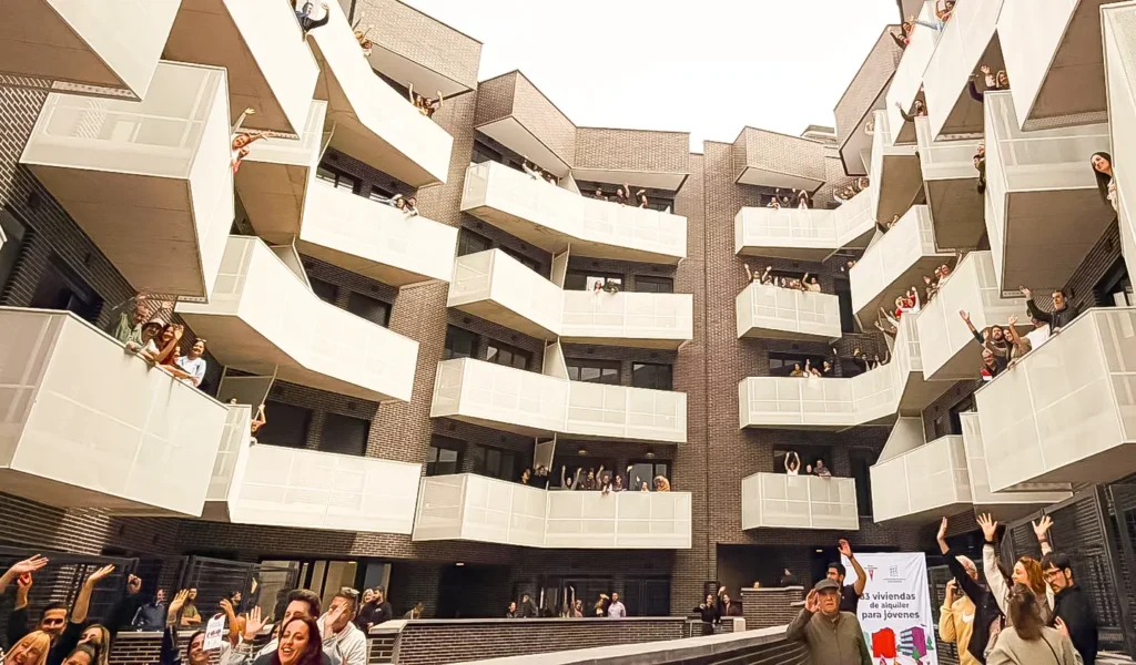 Un grupo de personas en varios pisos y balcones de un moderno complejo de apartamentos, parte de las viviendas públicas del Ayuntamiento de Rivas, saludan hacia la cámara, creando un ambiente animado y acogedor en la zona del patio.