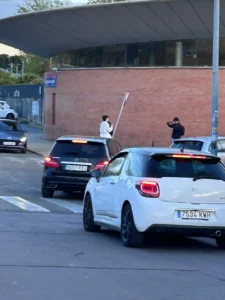 Un hombre con camisa blanca se encuentra en la acera sosteniendo una señal de tráfico cerca de un mural de sanidad pública, mientras otra persona le hace una foto. Varios coches esperan en un paso de peatones frente al edificio de ladrillo.
