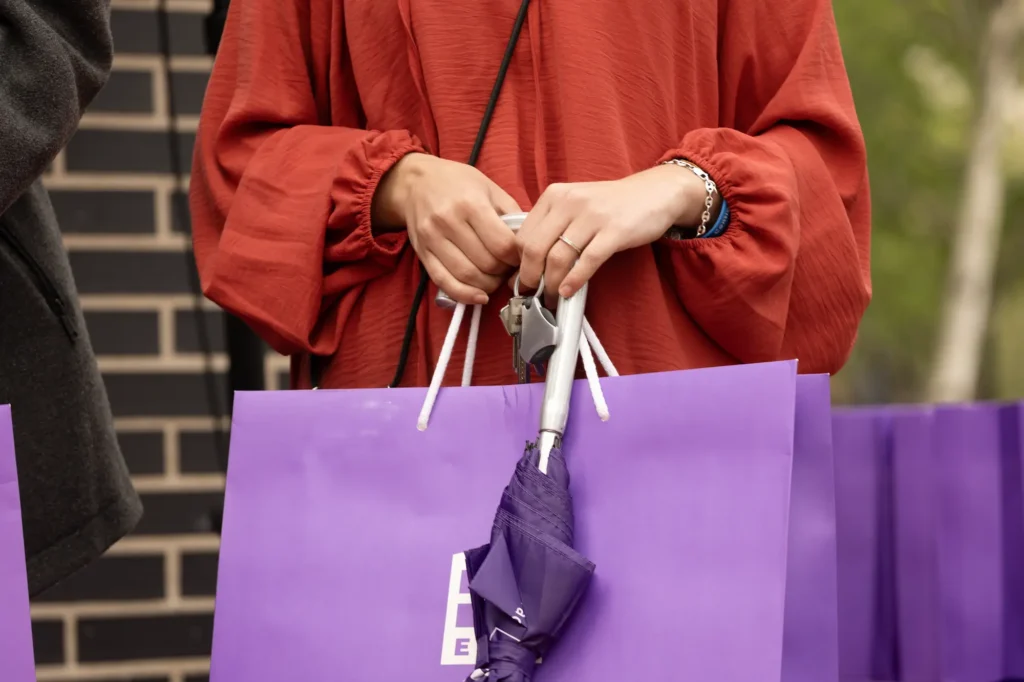 Una persona con camiseta roja de manga larga sostiene varias bolsas de la compra moradas y un paraguas morado doblado, junto a un muro de ladrillo y vegetación, quizá de camino a explorar las viviendas públicas promovidas por el Ayuntamiento de Rivas.