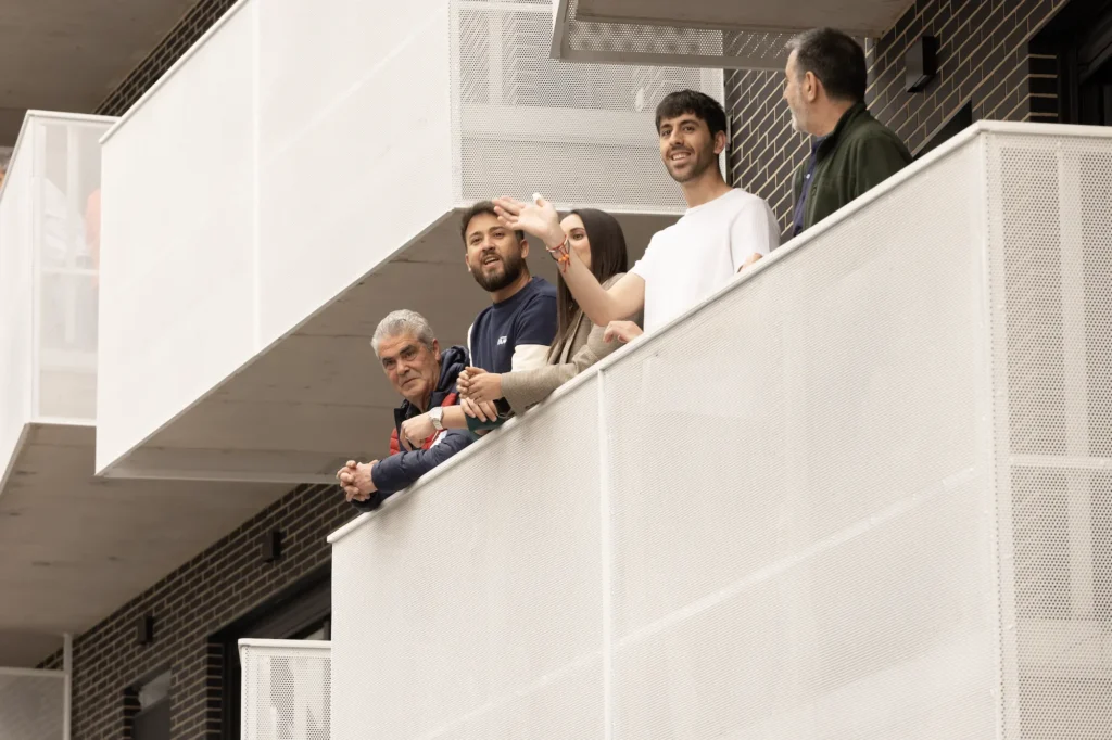 Cinco personas se asoman a un moderno balcón blanco con barandilla de rejilla en un bloque de viviendas públicas del Ayuntamiento de Rivas. Dos saludan y sonríen, mientras los otros miran al frente. El fondo muestra un edificio de ladrillo y otros balcones.